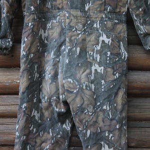 Puede incluir: Mono de camuflaje en tonos marrones, verdes y blancos. La prenda de una pieza presenta un estampado de hojas, mangas largas y un bolsillo trasero. El mono est&aacute; hecho de una tela duradera, adecuada para actividades al aire libre.