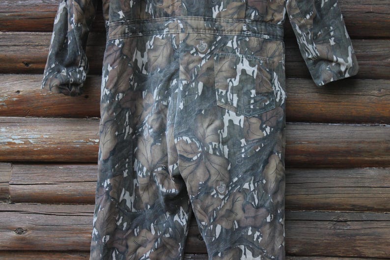 Puede incluir: Mono de camuflaje en tonos marrones, verdes y blancos. La prenda de una pieza presenta un estampado de hojas, mangas largas y un bolsillo trasero. El mono est&aacute; hecho de una tela duradera, adecuada para actividades al aire libre.