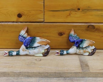 May include: Two ceramic duck figurines with outstretched wings, painted in shades of blue, green, brown, and white. The ducks are lying down, with their beaks open. The figurines are on a wooden surface.