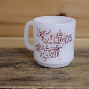 Peut inclure: Une tasse à café en céramique blanche avec un contour rouge du texte "La Musique et Moi" et des étoiles. La tasse a une poignée et est posée sur une surface en bois.