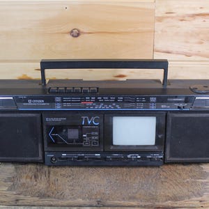 May include: A vintage black Citizen boombox with a built-in TV screen. The boombox has a handle, speakers on either side, and a cassette player. The TV screen is white, and the boombox is on a wooden surface.
