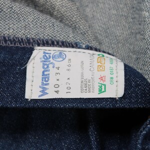 May include: A blue denim label with white text that reads "Wrangler" and "40 x 34". The label also includes the text "102 x 86 cm", "COTTON 100% COTON", "CA 00121", "MADE IN FABRIQUE AU CANADA", and "OBW 0187 400".