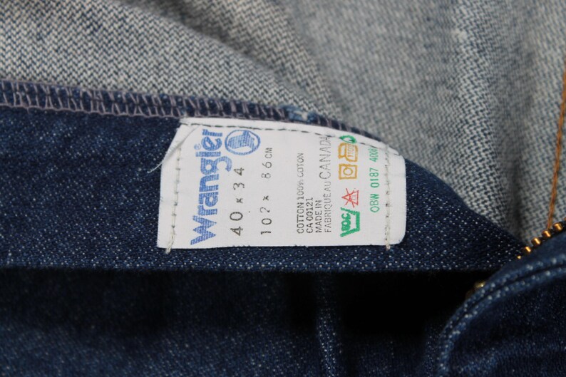 May include: A blue denim label with white text that reads "Wrangler" and "40 x 34". The label also includes the text "102 x 86 cm", "COTTON 100% COTON", "CA 00121", "MADE IN FABRIQUE AU CANADA", and "OBW 0187 400".