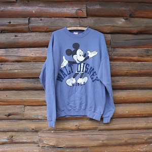 Puede incluir: Una sudadera azul b&iacute;garo con un gr&aacute;fico de Mickey Mouse y el texto "WALT DISNEY WORLD" en blanco. La sudadera est&aacute; colgada de una percha met&aacute;lica sobre un fondo de madera.