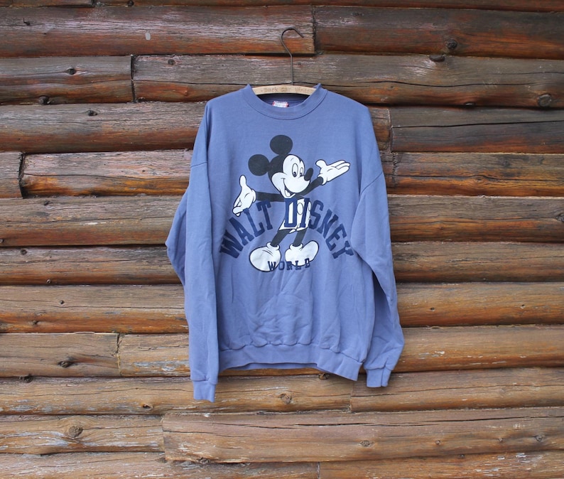 Puede incluir: Una sudadera azul b&iacute;garo con un gr&aacute;fico de Mickey Mouse y el texto "WALT DISNEY WORLD" en blanco. La sudadera est&aacute; colgada de una percha met&aacute;lica sobre un fondo de madera.