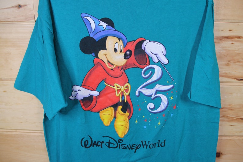 May include: A teal blue t-shirt featuring a graphic of Mickey Mouse dressed as a sorcerer. Mickey is holding a wand and pointing it at the number 25. The text "Walt Disney World" is printed below the graphic.
