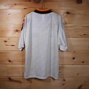 May include: White soccer jersey with yellow and black trim around the collar and sleeves. The jersey has a textured pattern and is made of a lightweight material.