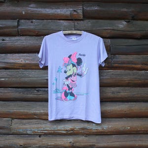 May include: A light purple short-sleeve t-shirt featuring a graphic of Minnie Mouse wearing a pink bow and sunglasses. The design includes the text Florida, palm trees, and a beach scene in shades of pink, blue, and black on the front center.