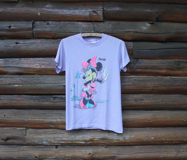 May include: A light purple short-sleeve t-shirt featuring a graphic of Minnie Mouse wearing a pink bow and sunglasses. The design includes the text Florida, palm trees, and a beach scene in shades of pink, blue, and black on the front center.