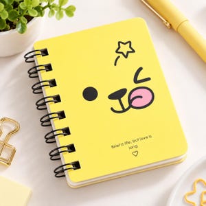 May include: A bright yellow spiral-bound notebook featuring a cartoon face with a black eye, a nose, and a pink tongue. The cover includes the text "Brief is life, but love is long" followed by a small heart symbol printed in black ink.