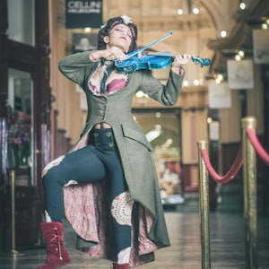 Puede incluir: Una persona con un atuendo de estilo vintage toca un violín azul. El conjunto incluye un abrigo largo verde, leggings oscuros con detalles de encaje y botas burdeos. La escena se desarrolla en un pasillo arqueado y ornamentado.