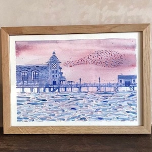 May include: Watercolor painting of a pier with buildings and a cloudy sky. The pier is blue, and the buildings are a mix of blue and white. The sky is pink and purple. The painting is framed in a light brown wooden frame.