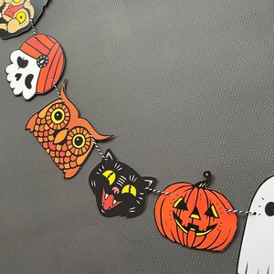 May include: A Halloween banner with orange, black, and white paper cutouts of an owl, a cat, a pumpkin, and a ghost. The banner is strung together with twine.