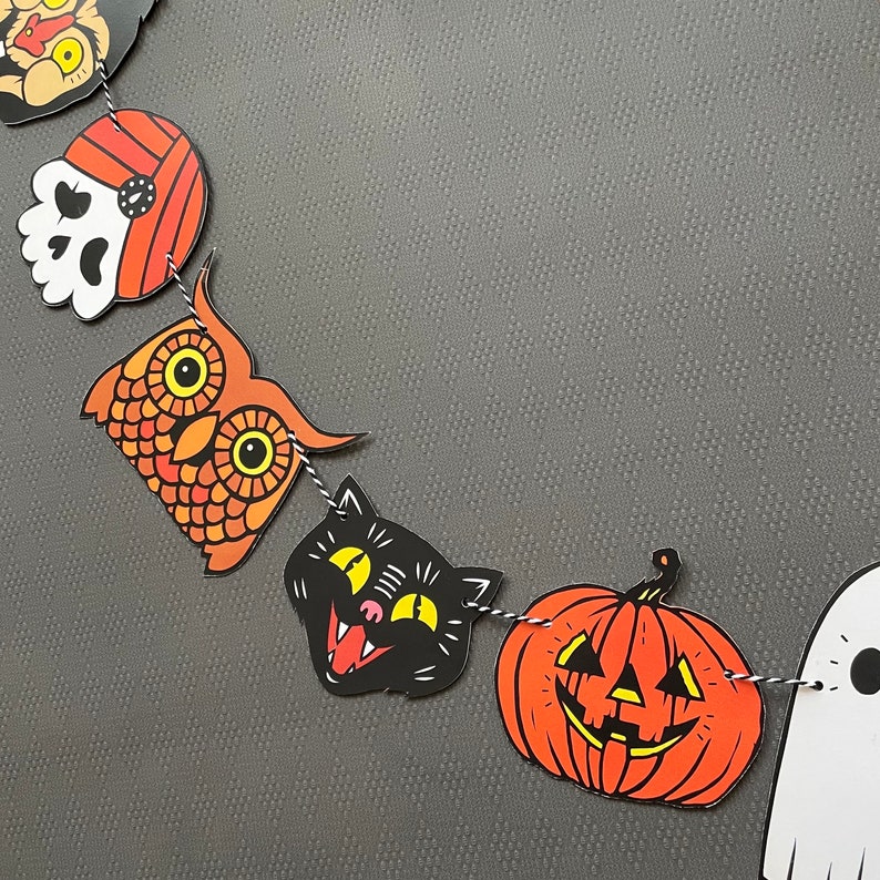 May include: A Halloween banner with orange, black, and white paper cutouts of an owl, a cat, a pumpkin, and a ghost. The banner is strung together with twine.