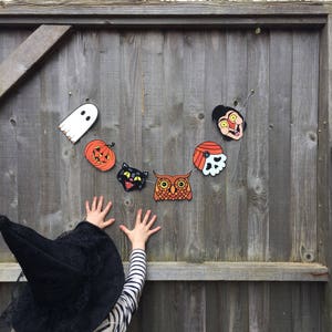 May include: A child wearing a black witch hat reaches up to a wooden fence decorated with a Halloween garland. The garland features a ghost, pumpkin, cat, owl, skull, and a character with a red nose and yellow eyes.