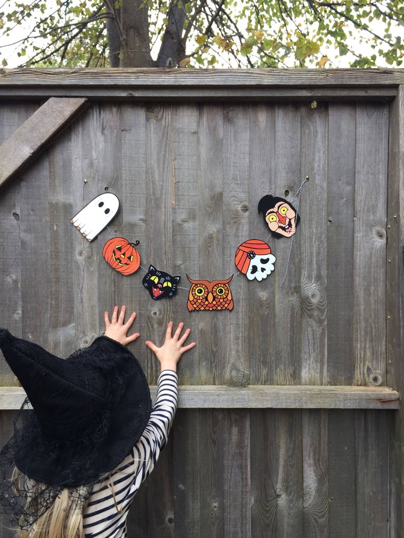 May include: A child wearing a black witch hat reaches up to a wooden fence decorated with a Halloween garland. The garland features a ghost, pumpkin, cat, owl, skull, and a character with a red nose and yellow eyes.