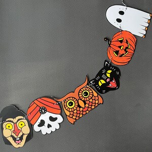 May include: A Halloween garland with orange, black, and white paper cutouts. The garland features a ghost, a pumpkin, a black cat, an owl, a skull, and a witch.