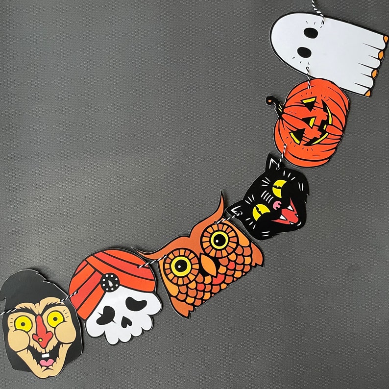 May include: A Halloween garland with orange, black, and white paper cutouts. The garland features a ghost, a pumpkin, a black cat, an owl, a skull, and a witch.