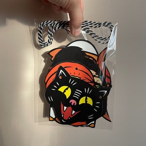 May include: A set of three Halloween-themed paper cutouts featuring a black cat, a pumpkin, and a ghost. The cutouts are strung together with black and white twine and are ready to be hung for Halloween decorations.