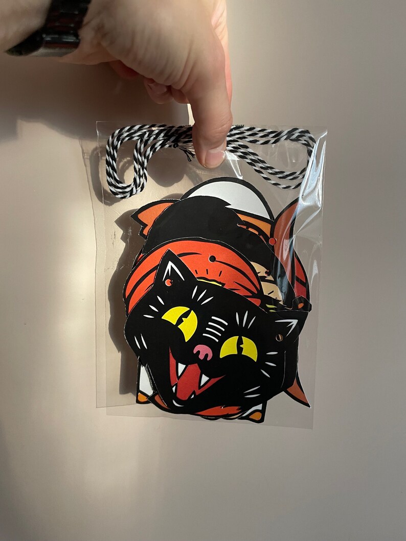 May include: A set of three Halloween-themed paper cutouts featuring a black cat, a pumpkin, and a ghost. The cutouts are strung together with black and white twine and are ready to be hung for Halloween decorations.