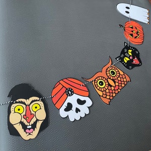 May include: A Halloween banner with six different characters. The characters are a ghost, a pumpkin, a black cat, an owl, a skull with a turban, and a witch. The banner is made of paper and is strung together with twine.