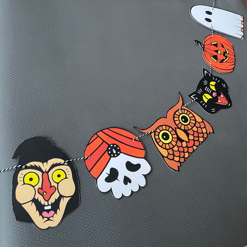 May include: A Halloween banner with six different characters. The characters are a ghost, a pumpkin, a black cat, an owl, a skull with a turban, and a witch. The banner is made of paper and is strung together with twine.