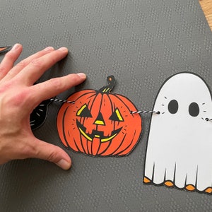 May include: A Halloween garland with a black cat, a jack-o'-lantern, and a white ghost. The garland is strung with black and white twine.