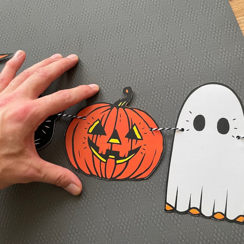 May include: A Halloween garland with a black cat, a jack-o'-lantern, and a white ghost. The garland is strung with black and white twine.