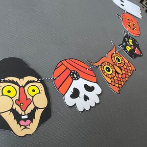 May include: A Halloween banner with a black and white skull, a red and orange turban, an orange owl, a black cat, a pumpkin, and a white ghost. The banner is strung together with black and white string.