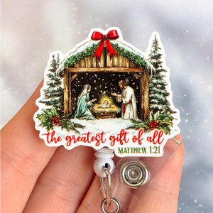 May include: A Christmas-themed badge reel featuring a Nativity scene with Mary, Joseph, and baby Jesus. The design includes a red bow, evergreen boughs, and the text "the greatest gift of all" with a Bible verse reference.