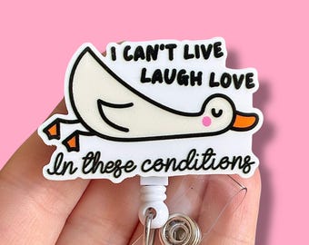 May include: White plastic badge holder with a cartoon duck design. The duck is upside down and the text reads "I can't live laugh love in these conditions".