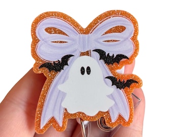 May include: A Halloween-themed badge reel featuring a white ghost with black eyes and two black bats. The ghost is set against a white bow and an orange glitter background. The badge reel has a retractable cord and a metal clip.