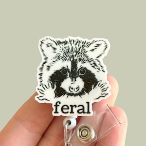 May include: A white badge holder with a black and white illustration of a raccoon peeking out from behind the text "feral".