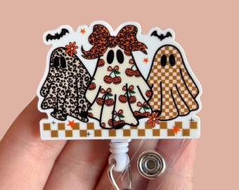 May include: A Halloween-themed badge reel featuring three cartoon ghosts with unique patterns. One ghost has a leopard print, another has a cherry pattern, and the third has a checkered design. The badge reel has a white base with a decorative border.
