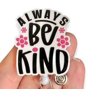May include: A white and black badge reel with the words "ALWAYS BE KIND" in a bold, playful font. Pink flower accents surround the text. The badge reel has a clear plastic clip and a retractable cord.