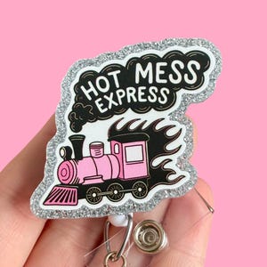 May include: A silver glitter badge holder with a pink and black cartoon train and the text "Hot Mess Express".