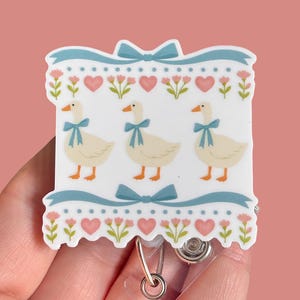 May include: A white badge reel featuring three geese with blue bows, set against a backdrop of pink hearts and flowers. The design is framed by a blue ribbon with bows, creating a charming, whimsical aesthetic.