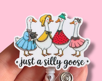 May include: A white badge holder with a clear plastic retractable reel. The front of the badge holder features a cartoon image of four geese wearing colorful dresses and hats. The text "just a silly goose" is printed below the geese.