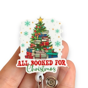 May include: A white Christmas tree-shaped badge holder with a green Christmas tree decorated with red and gold ornaments and a stack of books. The text "All Booked for Christmas" is printed below the tree.