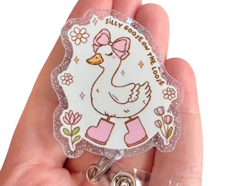 May include: A badge reel with a cartoon goose wearing a pink bow and boots, surrounded by flowers and the text "Silly Goose on the Loose." The badge has a glittery border and a clear plastic clip.
