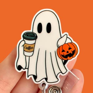 May include: A white ghost holding a coffee cup that says "Coffee" and a small orange jack-o'-lantern. The ghost is attached to a clear plastic retractable badge holder.