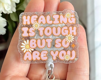 May include: A clear glitter badge holder with a pink and white design. The badge holder has the text "Healing is tough but so are you" with daisy flowers.