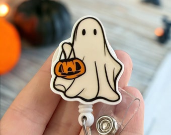 May include: White ghost with a friendly face holding a small orange pumpkin. The ghost is attached to a clear plastic retractable badge holder with a silver clip.