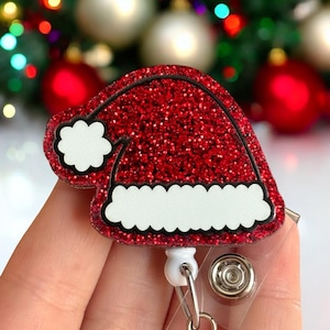 May include: Red glitter Christmas Santa hat badge reel with a white pom pom and white trim. The reel is attached to a clear plastic badge holder.