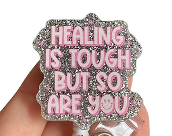 May include: Silver glitter badge holder with a pink glitter background and the text "HEALING IS TOUGH BUT SO ARE YOU" with a pink smiley face.