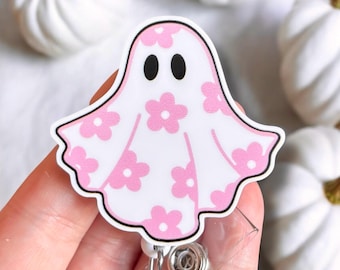 May include: A white and pink floral ghost badge reel. The ghost has black eyes and is decorated with pink flowers. The badge reel has a clear plastic strap and a metal clip.