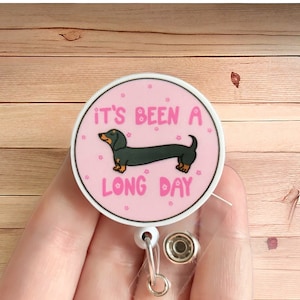 May include: Pink round badge holder with a black dachshund and the text "It's been a long day".