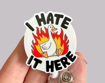 May include: White retractable badge holder with a round, white plastic charm featuring a cartoon goose in flames with the text "I hate it here".