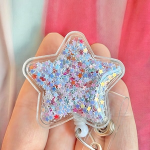 K&ouml;nnte beinhalten: Ein durchsichtiger, sternf&ouml;rmiger Badgehalter aus Plastik, gef&uuml;llt mit schimmerndem Glitzer in Blau-, Rosa- und Goldt&ouml;nen. Der Halter ist an einer einziehbaren Badge-Spule mit einem silbernen Clip befestigt.