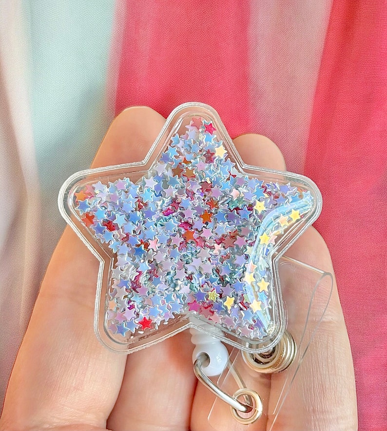 K&ouml;nnte beinhalten: Ein durchsichtiger, sternf&ouml;rmiger Badgehalter aus Plastik, gef&uuml;llt mit schimmerndem Glitzer in Blau-, Rosa- und Goldt&ouml;nen. Der Halter ist an einer einziehbaren Badge-Spule mit einem silbernen Clip befestigt.